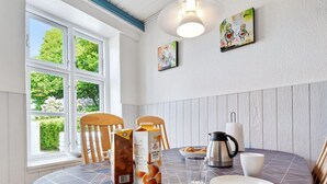 Uma área de refeições com uma mesa posta para dois, com toalha de mesa estampada, cafeteira, copo e caixas de suco.