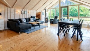Een ruime woonkamer met houten plafond, een zithoek, een open haard en een eettafel met stoelen.