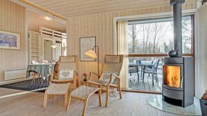 A cozy cabin interior with a wood-burning stove, wooden furniture, and a large window overlooking a snowy landscape.