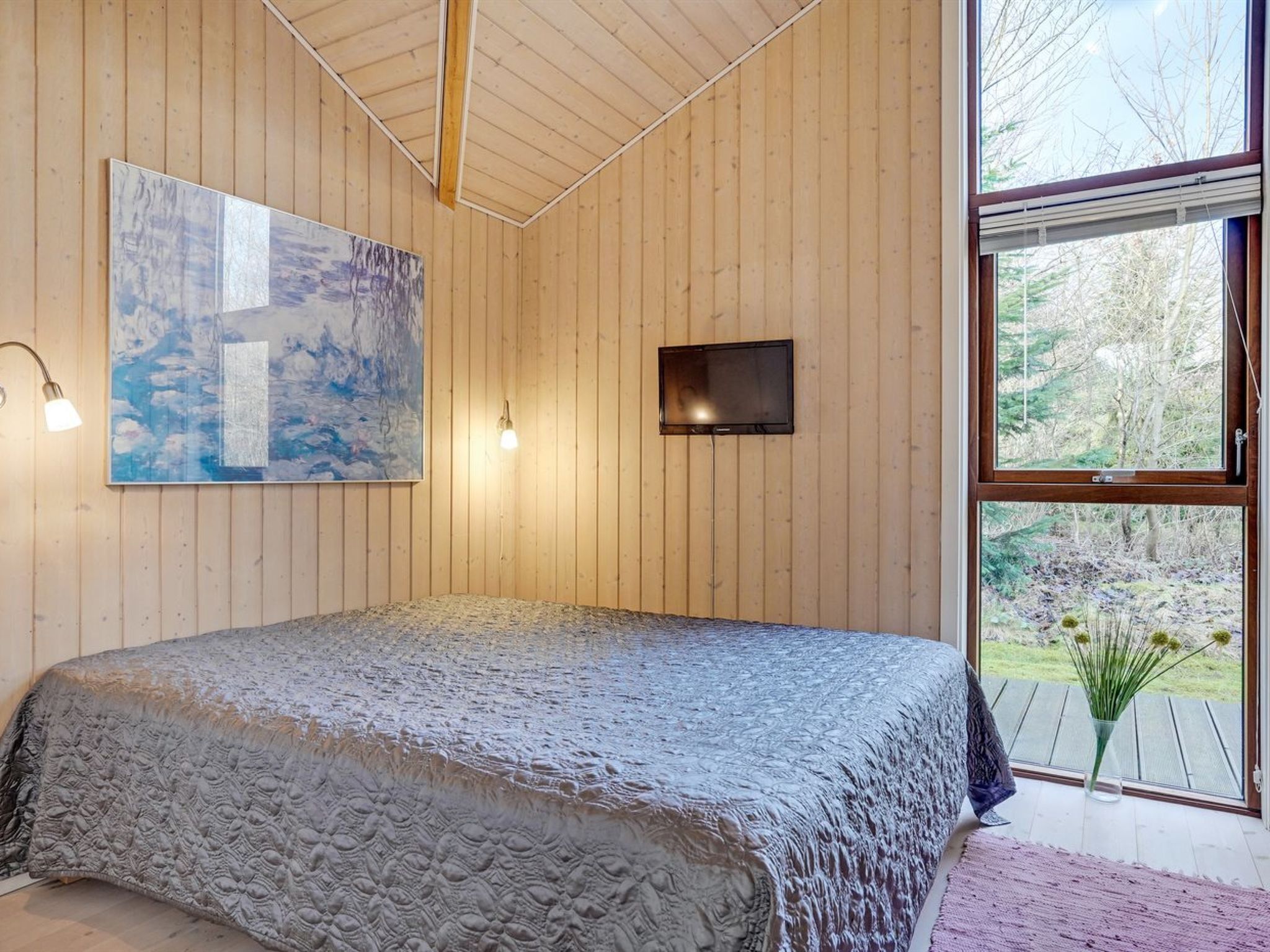 A bedroom with a large bed, wooden walls, a painting, a TV, and a large window with a view of trees.