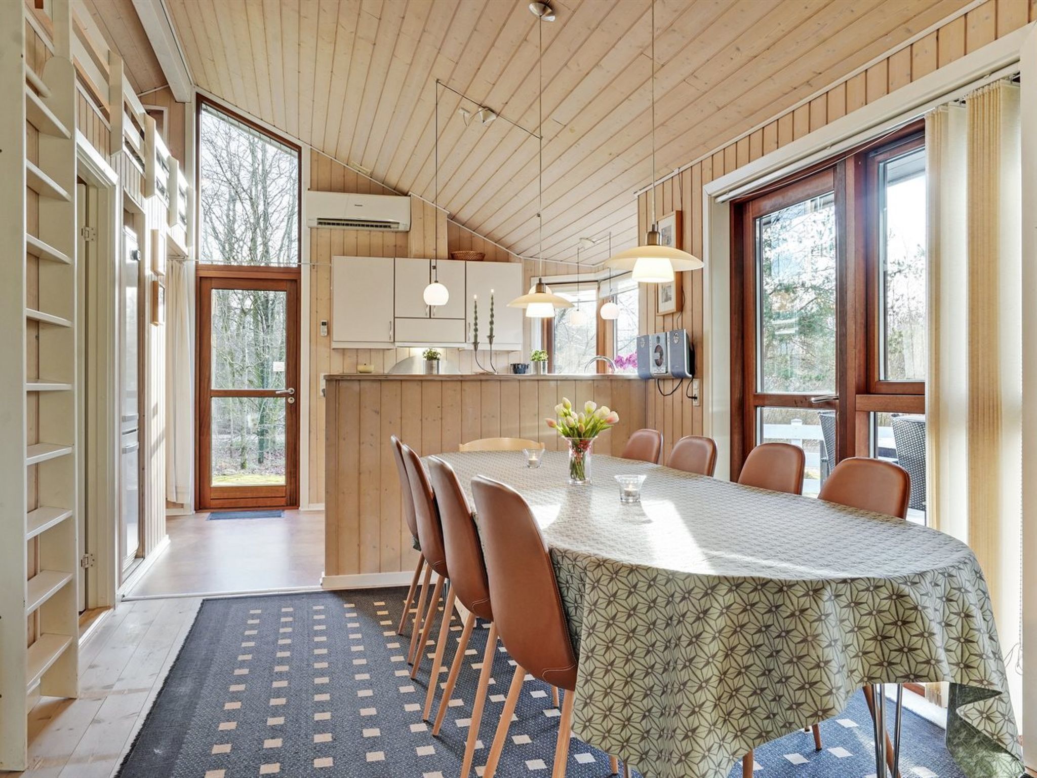 A dining area with a long table, chairs, and a patterned tablecloth. There are wooden cabinets and a window with a view of trees.