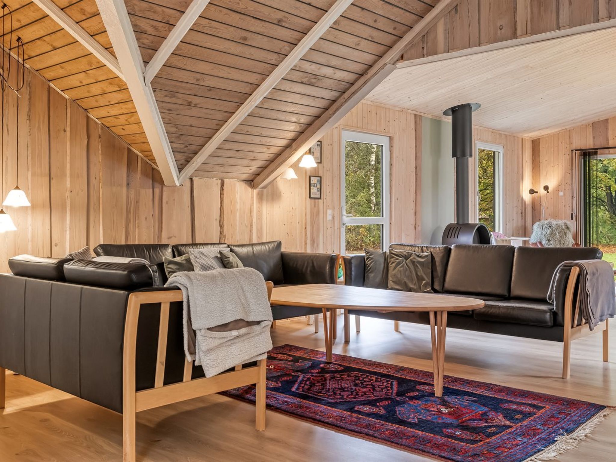 L’intérieur chaleureux d’un chalet avec un plafond en bois, un canapé en cuir noir, une table basse en bois et une grande fenêtre donnant sur le paysage extérieur.