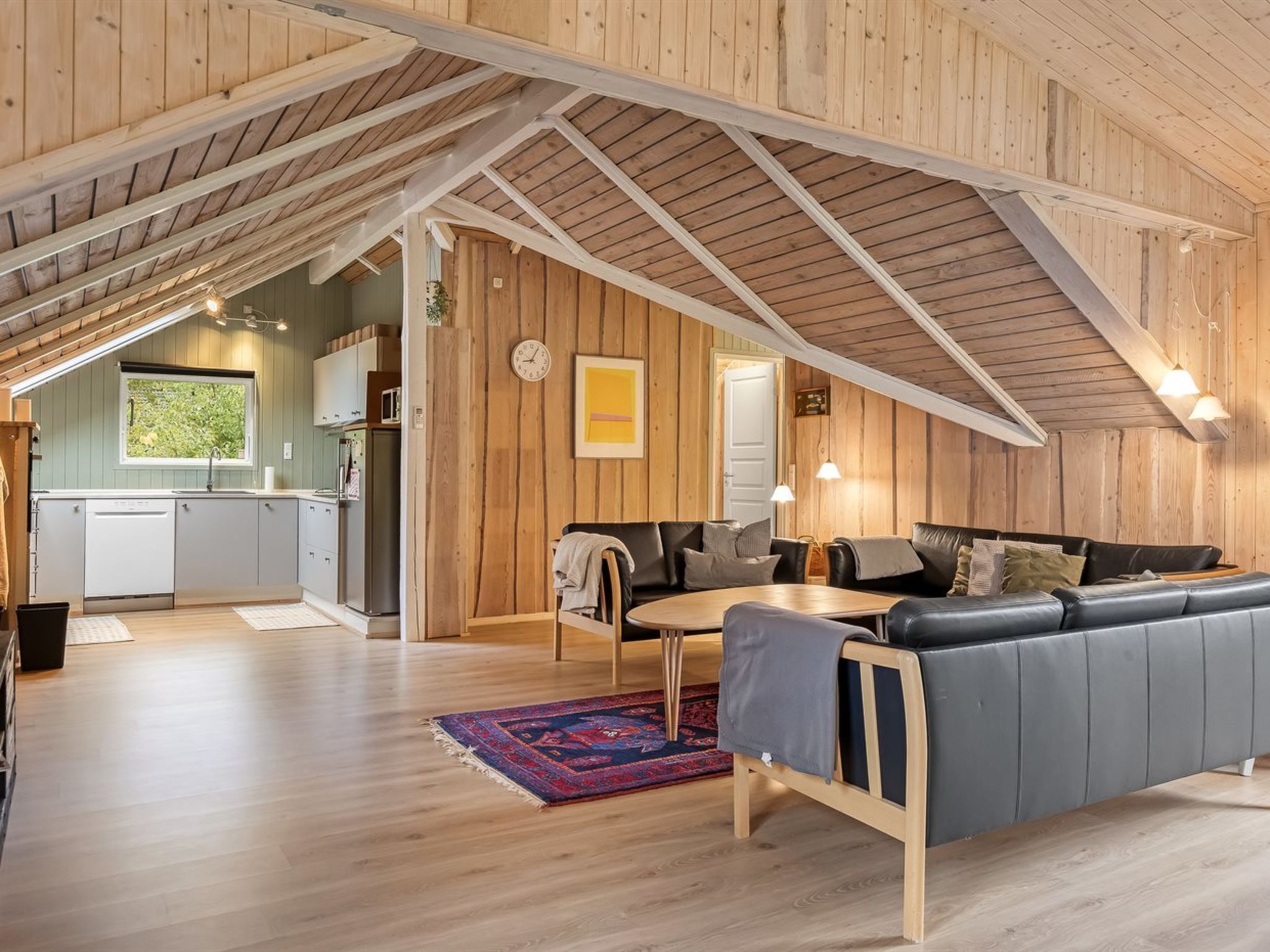 A spacious loft with a high ceiling, wooden beams, a kitchen with a window, and a cozy seating area with a rug.
