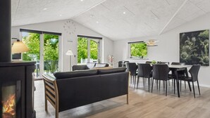 Una sala de estar moderna con chimenea, un sofá, un comedor con sillas y ventanas amplias con vistas a la vegetación.