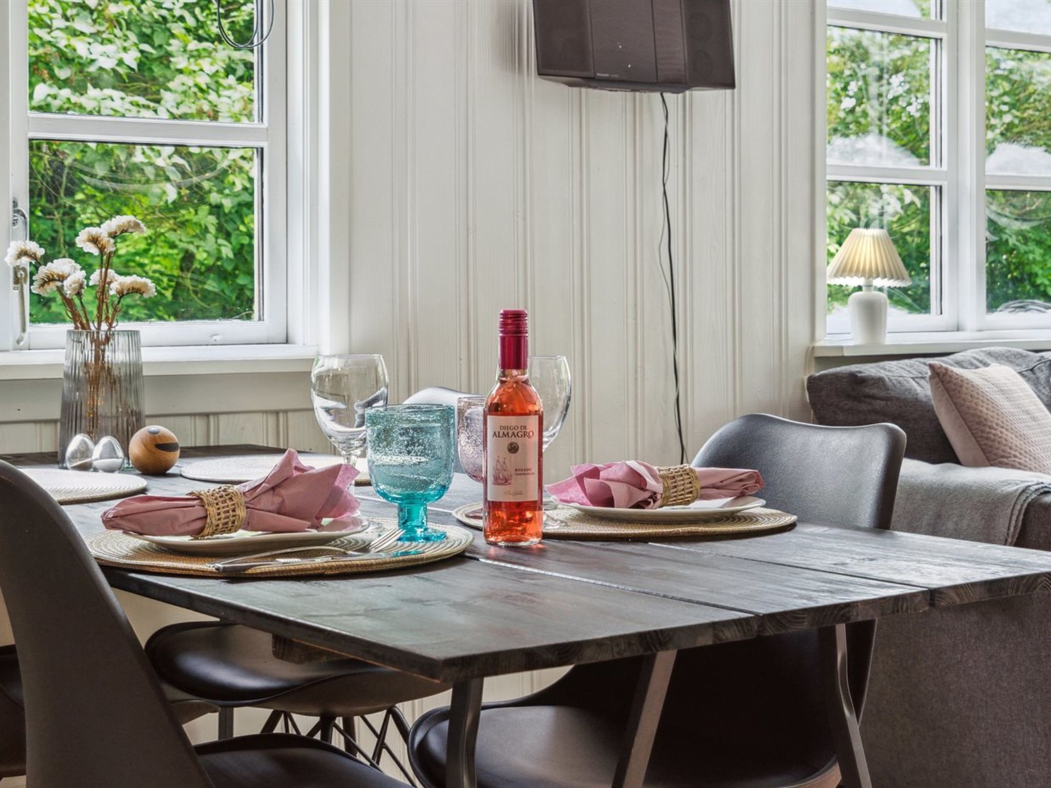 A dining table set with plates, glasses, and a bottle of wine, with a view of greenery outside.