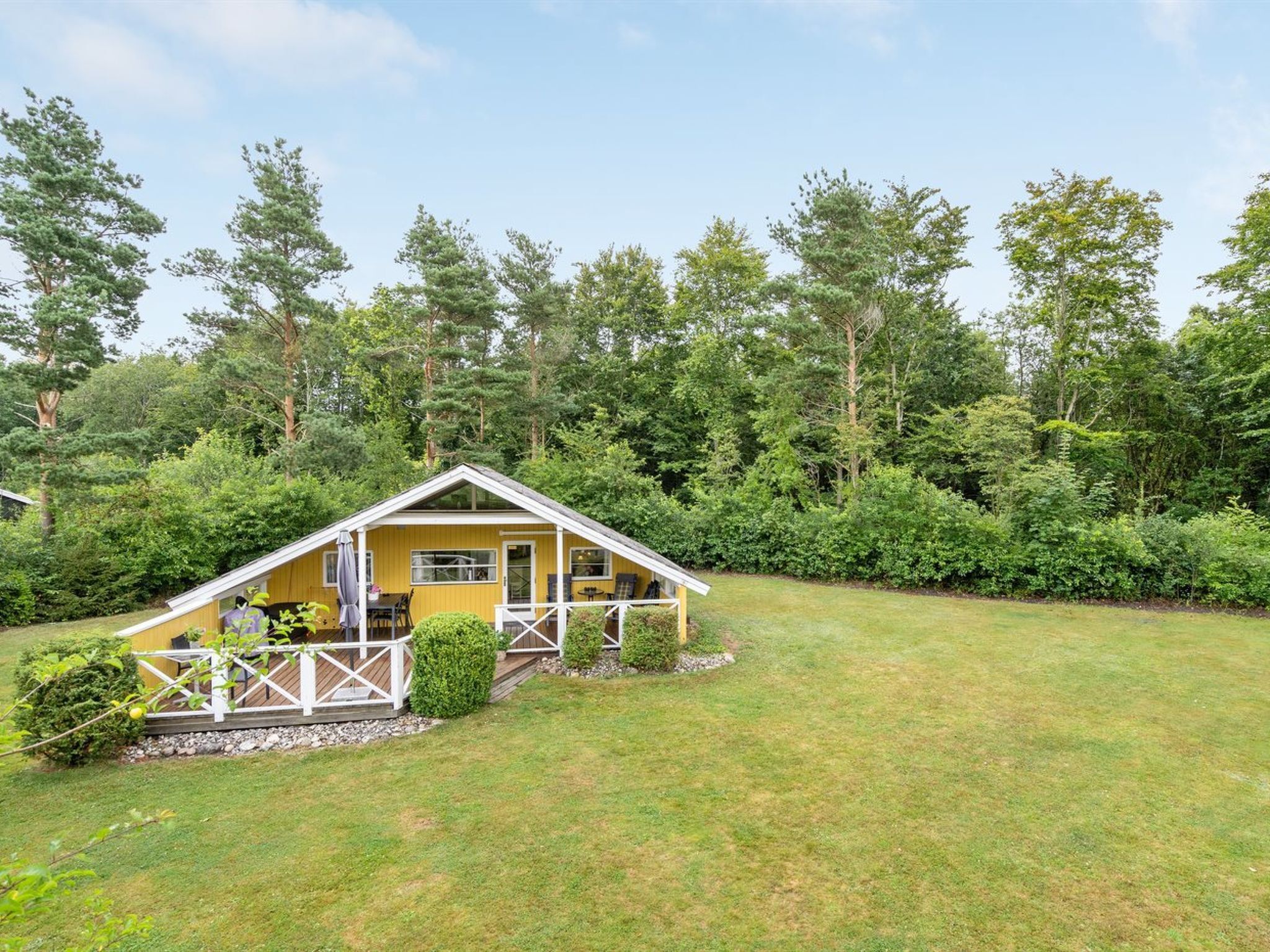 Une cabane jaune avec un porche, entourée d’une cour verdoyante et d’une forêt dense.