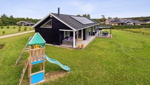 Een modern huis met een zwarte gevel, een overdekte patio en een speelruimte met een glijbaan.
