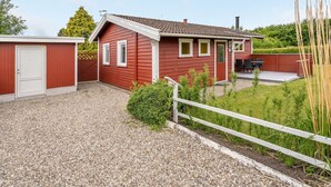Ein rotes Einfamilienhaus mit überdachter Terrasse, einem Schotterweg und einem weißen Zaun.