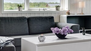 Una sala de estar moderna con un sofá gris, una mesa de centro blanca con un jarrón decorativo, y vistas a zonas verdes a través de grandes ventanales.