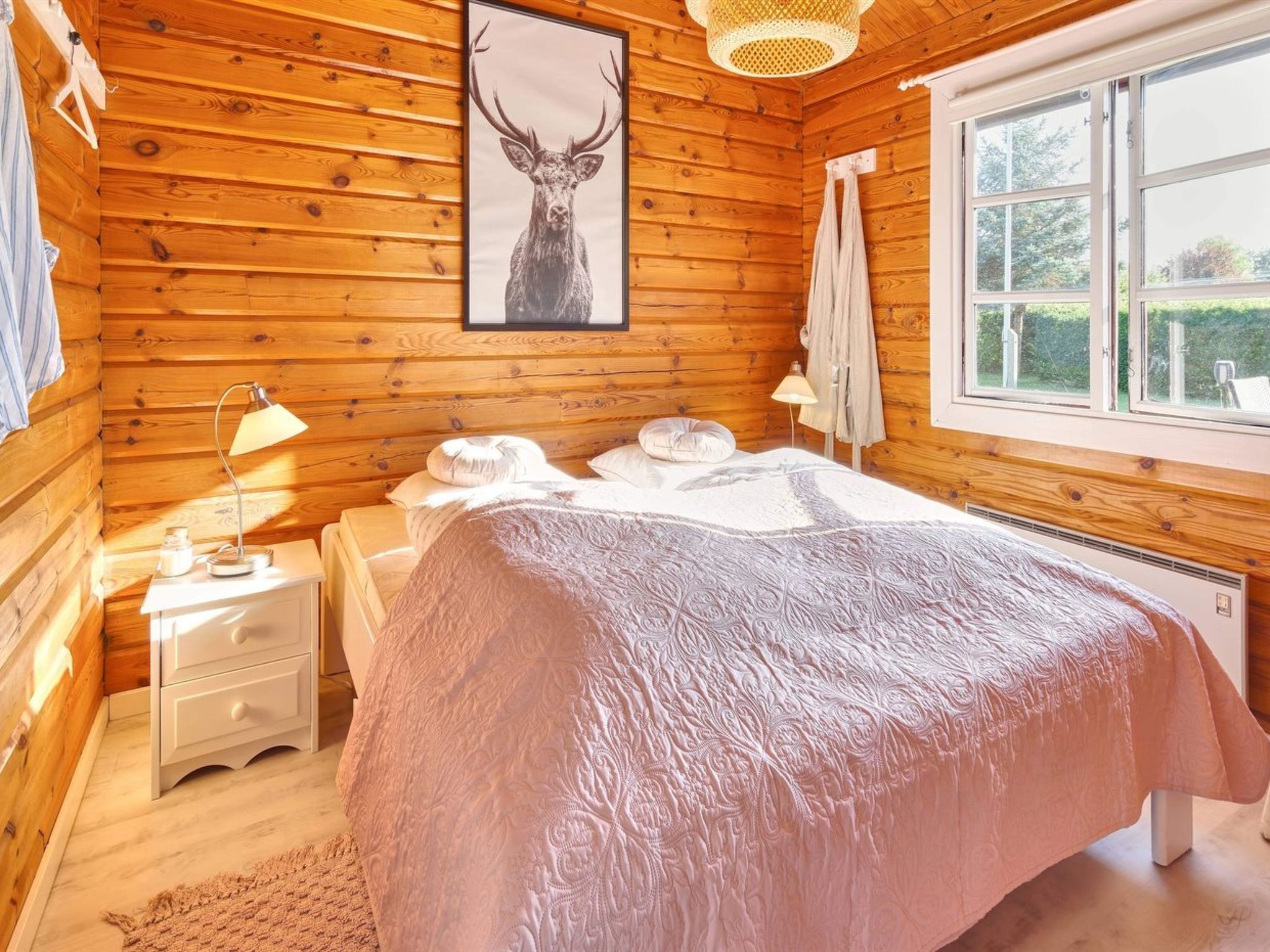 Une chambre confortable aménagée dans un chalet en bois, avec un grand lit, une table de chevet, une lampe et un tableau représentant un cerf accroché au mur.