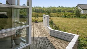 Una terraza de madera con barandilla blanca, una planta en maceta y vistas a un campo verde y árboles.