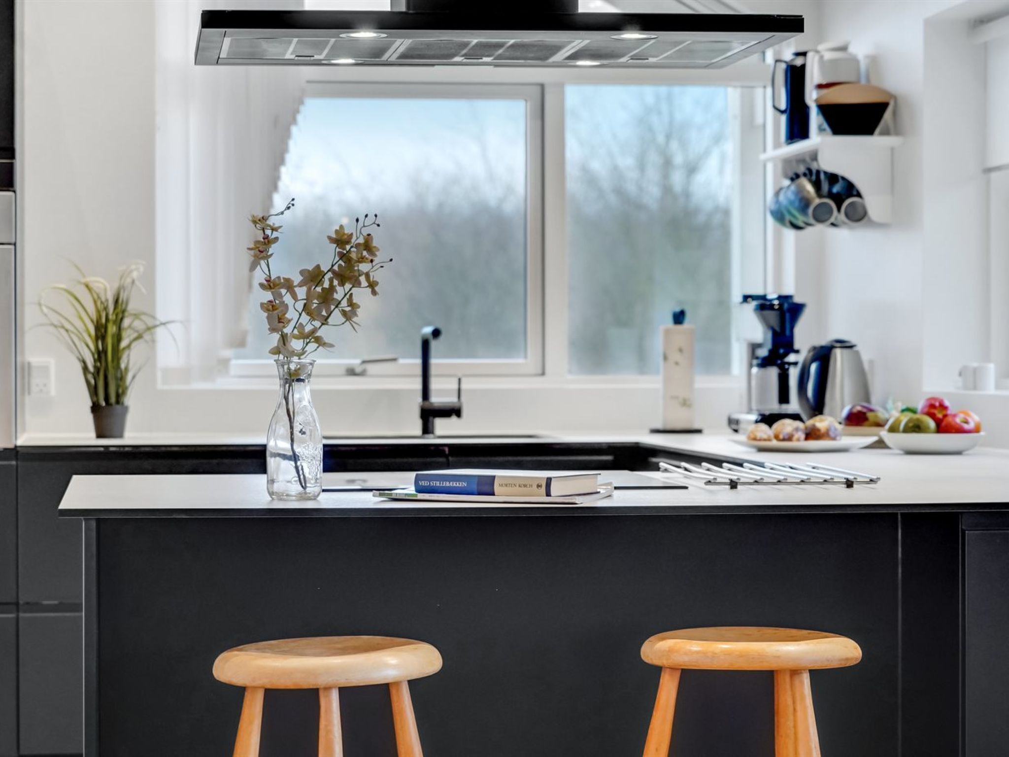 Une cuisine moderne avec un îlot central, deux tabourets en bois, un vase rempli de fleurs et une fenêtre donnant sur des arbres.