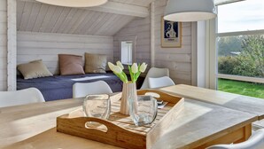 A cozy cabin bedroom with a wooden ceiling, a bed with pillows, a bedside table with a vase of flowers, and a window with a view of greenery.