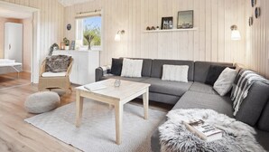 A modern living room with a grey sofa, a wooden coffee table, and a wicker chair. There is a window with a view of trees, and a shelf with decorative items.