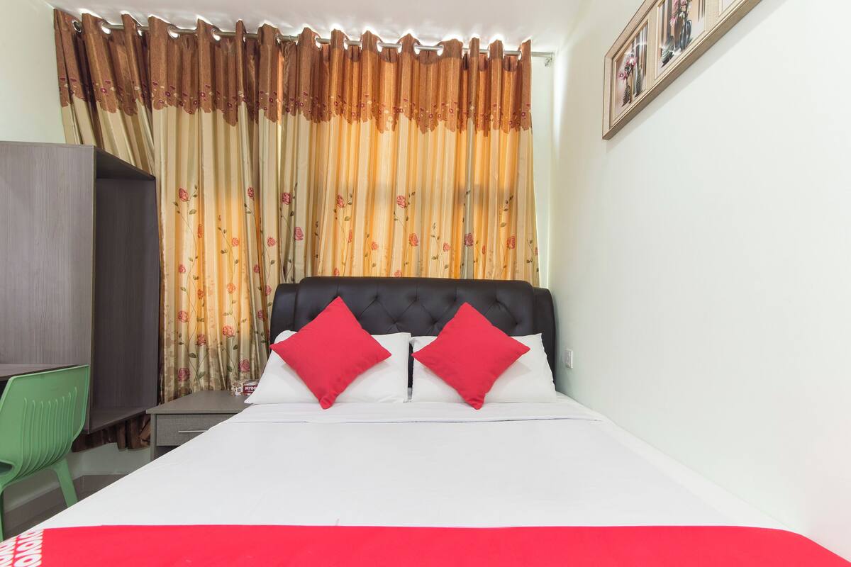 Quarto de hotel com cama, duas almofadas vermelhas, uma cadeira verde, escrivaninha e guarda-roupa.