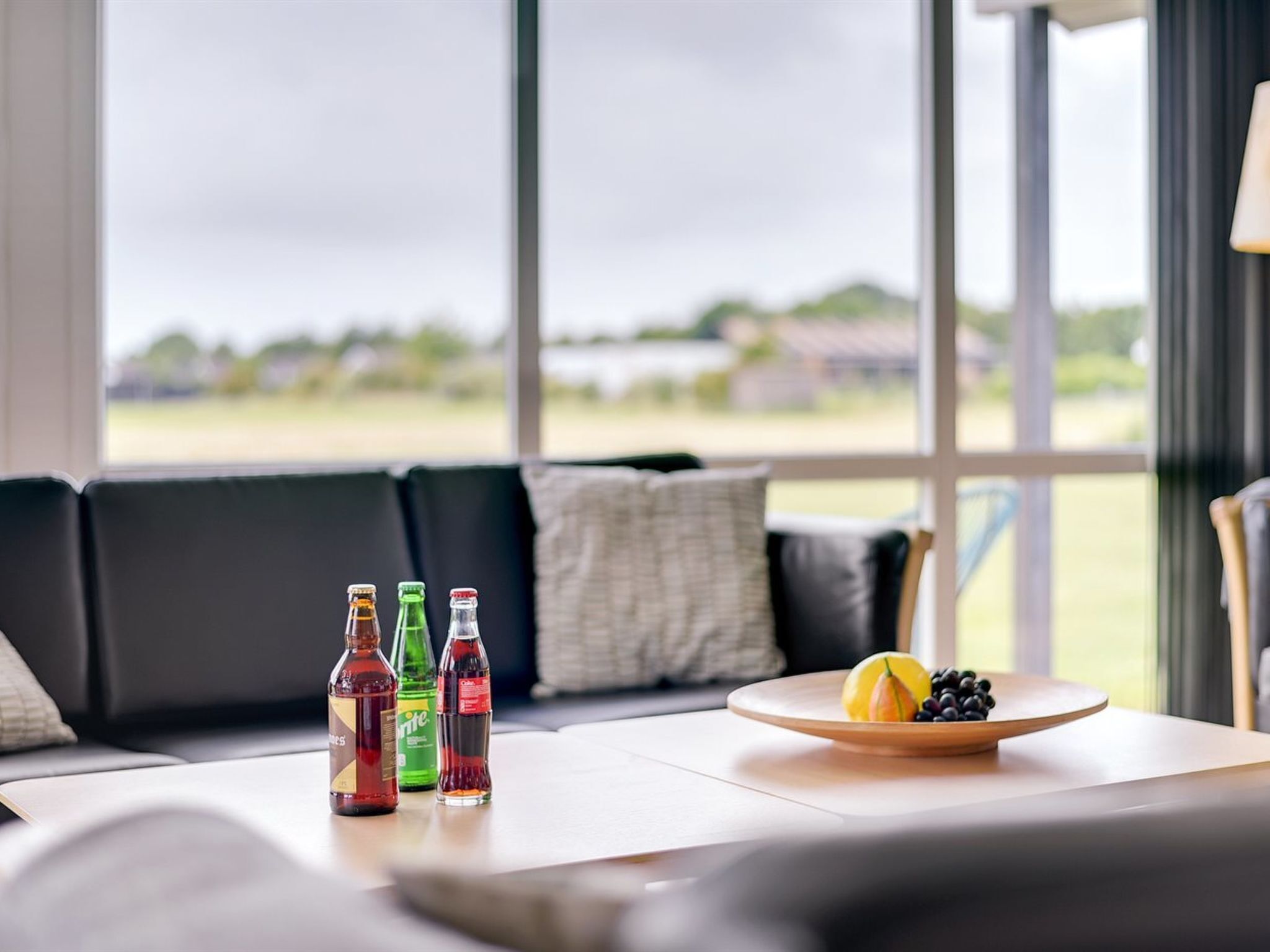 Een moderne woonkamer met een groot raam, een bank, een bijzettafeltje en een schaal fruit met flessen.