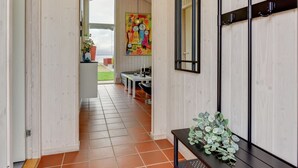 Een hal met houten wanden, een betegelde vloer, een bank, een tafel met een plant en een kleurrijk schilderij aan de muur.