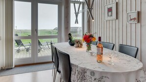 Ein Essbereich mit einem Tisch für zwei Personen, einer Blumenvase, einer Weinflasche und Blick ins Freie durch große Fenster.
