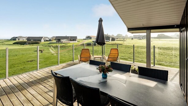 Ein überdachter Außenbereich mit Tisch und Stühlen, der auf ein grünes Feld und Gebäude in der Ferne blickt.