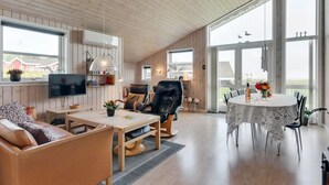 Ein großzügiger Wohnbereich mit Holzbalkendecke, einem Fernseher, einem Sofa, einem Esstisch und Blick durch große Fenster.