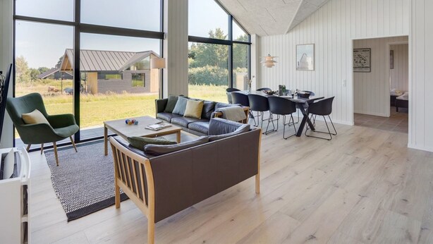 Een ruime woonkamer met een groot raam, een bank, een eettafel en een uitzicht op een huis op de achtergrond.