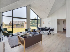 Een ruime woonkamer met een groot raam, een bank, een eettafel en een uitzicht op een huis op de achtergrond.