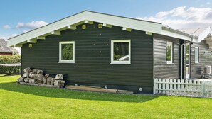 A single-story, dark green wooden house with a white picket fence, two windows, and a small porch area.