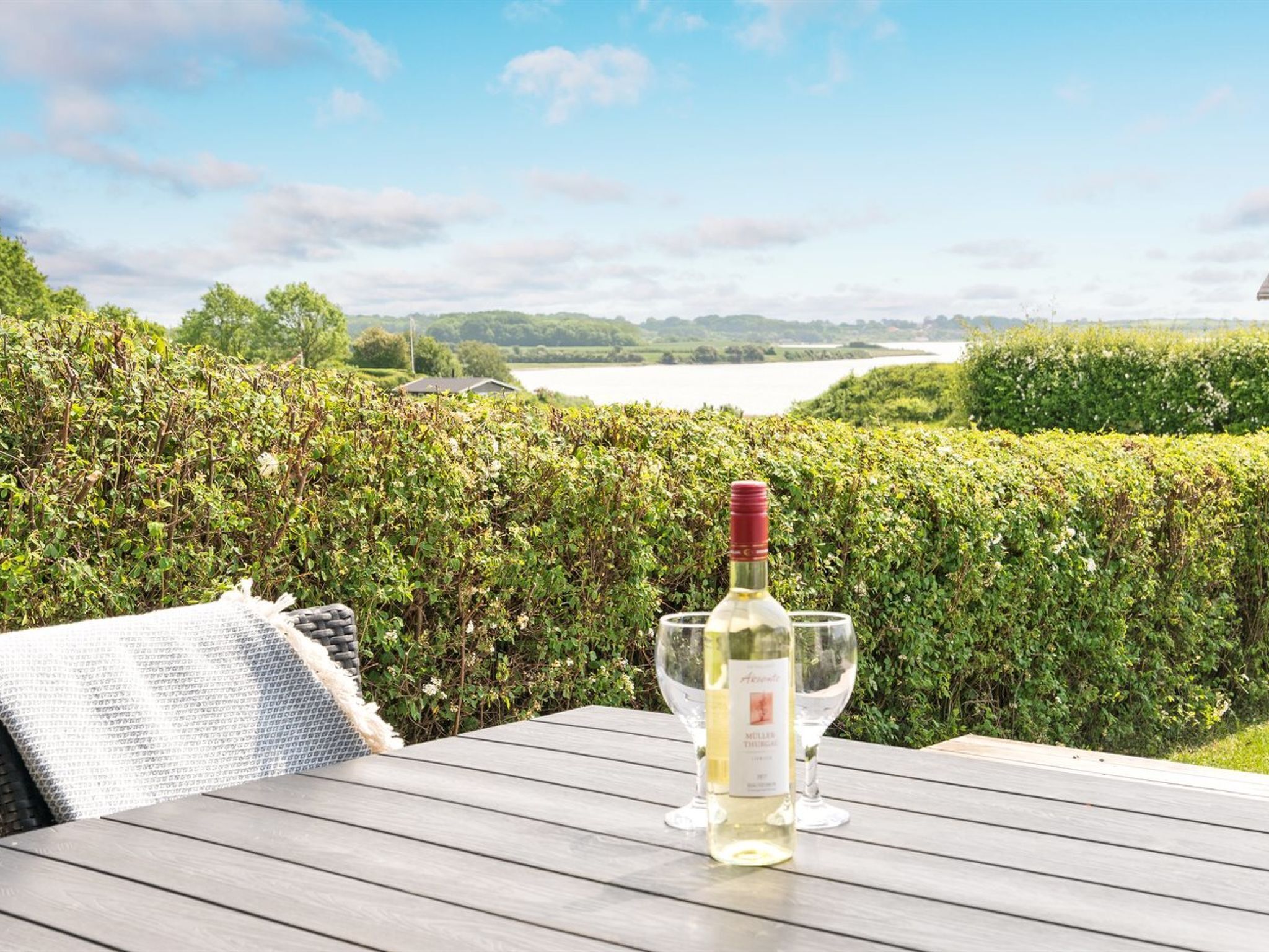 Une table avec une bouteille de vin et un verre, placée devant un jardin luxuriant et un lac.