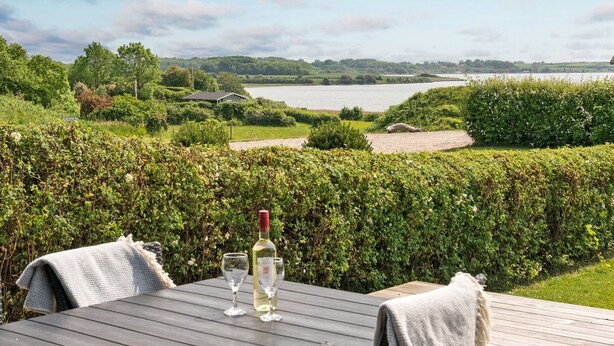 A table with a bottle of white wine and two glasses, overlooking a lake with a house and greenery in the background.