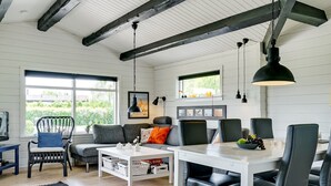 A modern living room with a white table, black chairs, and a sofa with grey cushions. There is a window with a view of greenery outside.