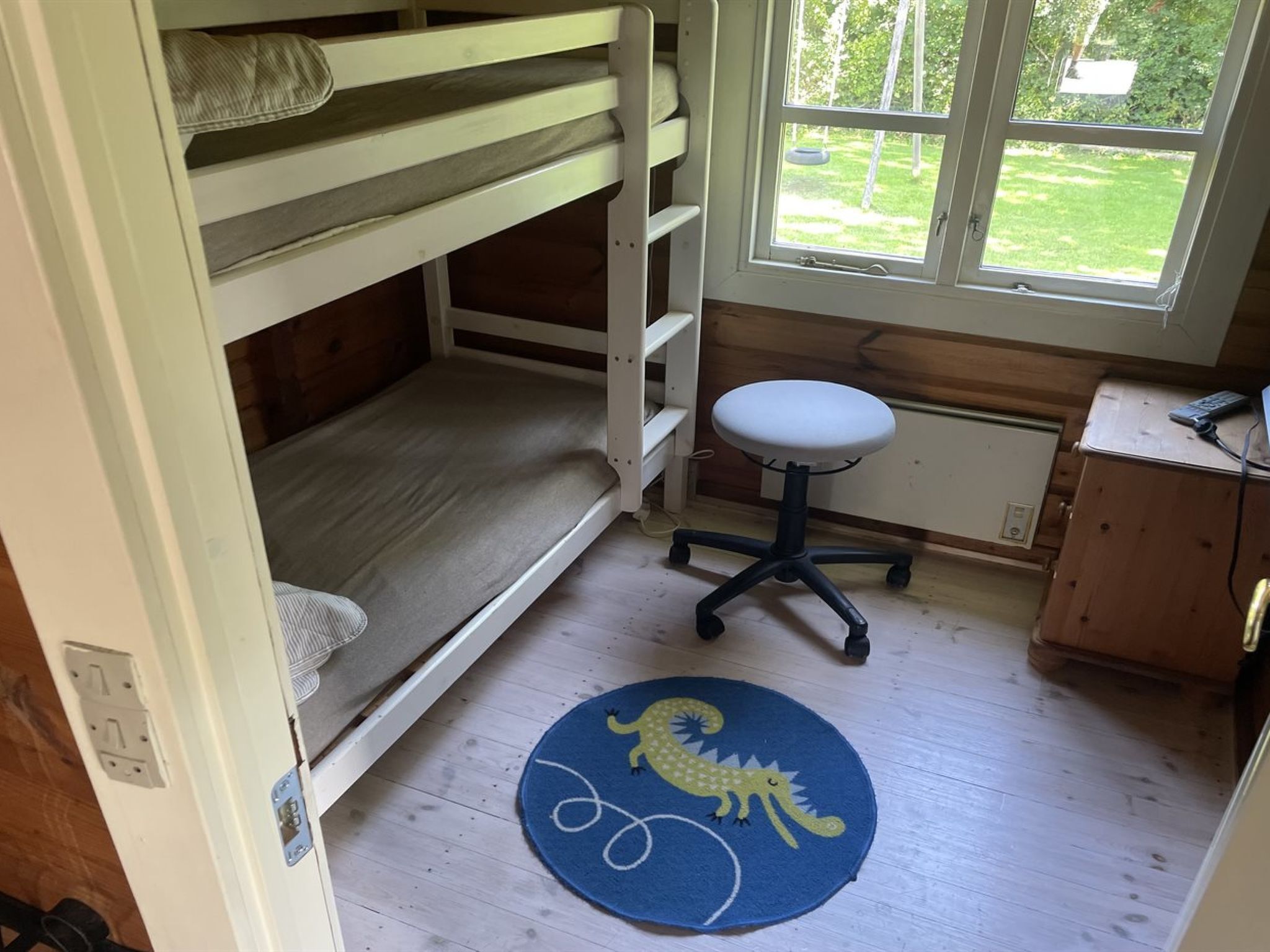 A bunk bed room with a desk and chair, a window with a view of greenery, and a blue rug with a lizard design.