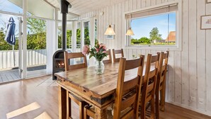 Une table à manger en bois avec des chaises, un vase rempli de fleurs, un poêle à bois et une fenêtre donnant sur un paysage verdoyant avec une maison.
