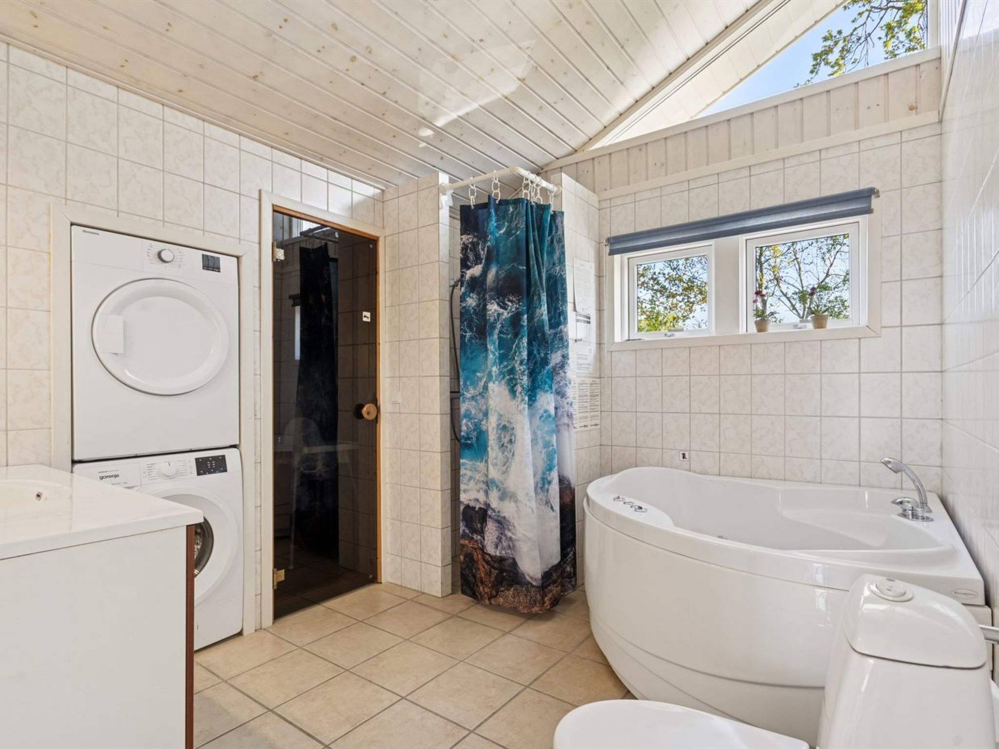Une salle de bain moderne équipée d’un lave-linge, d’une douche avec un rideau bleu en forme de cascade, d’une baignoire et d’une fenêtre donnant sur la verdure.