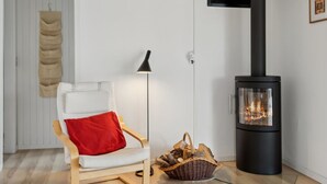 A modern living room with a white air conditioner, a black wood-burning stove, a white chair with a red pillow, a floor lamp, and a basket of firewood.