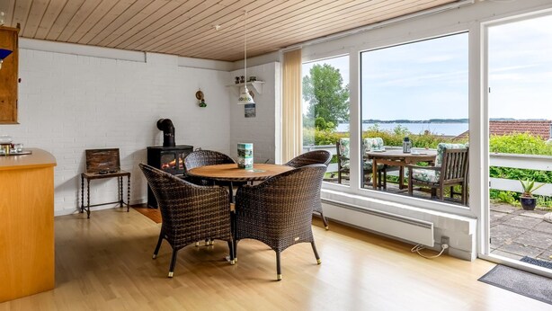 Ein gemütliches Zimmer mit Holzbalkendecke, einem runden Tisch mit Korbstühlen, einer kleinen Küchenzeile mit Herd und großen Fenstern mit Blick ins Freie.