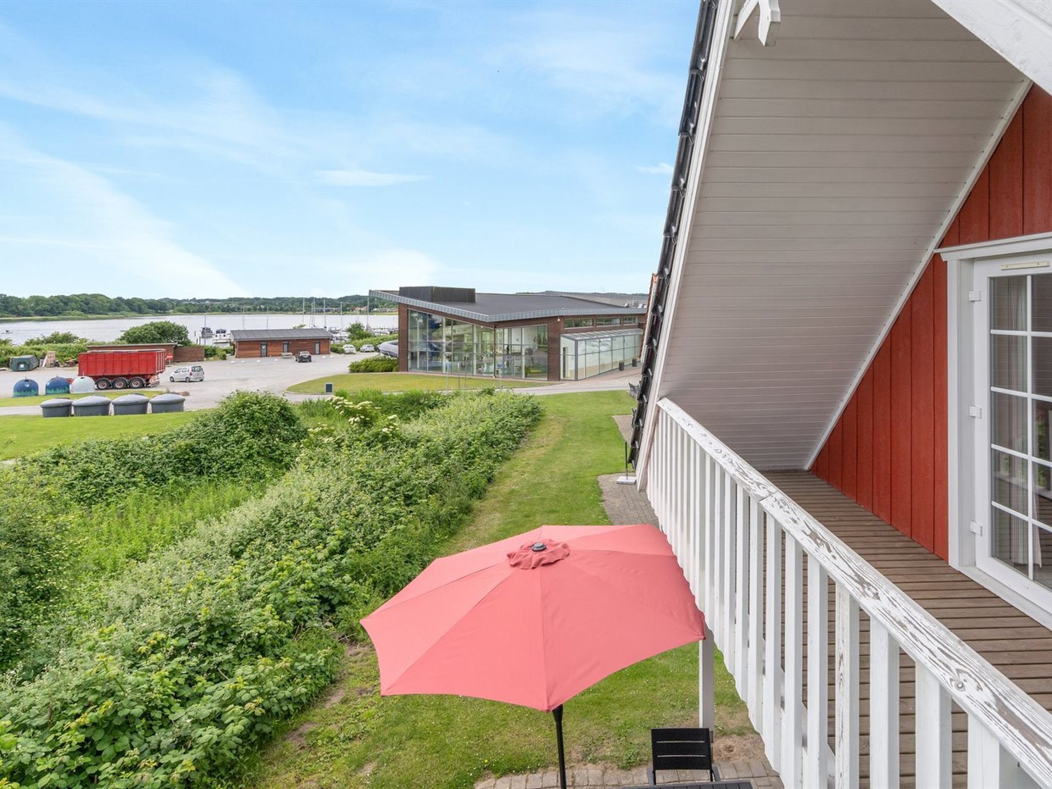 A building with a red exterior, a balcony with a red umbrella, and a view of a parking area and greenery.