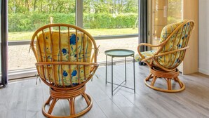 Ein Zimmer mit zwei Korbstühlen mit floralen Kissen, einem kleinen runden Tisch und Blick auf eine grüne Landschaft durch große Fenster.