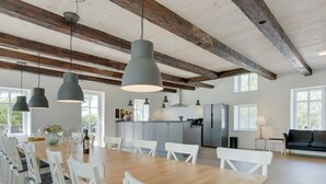 Un comedor amplio con una larga mesa de madera y sillas blancas, una cocina con armarios grises y un sofá de cuero negro en una habitación luminosa con vigas de madera en el techo.