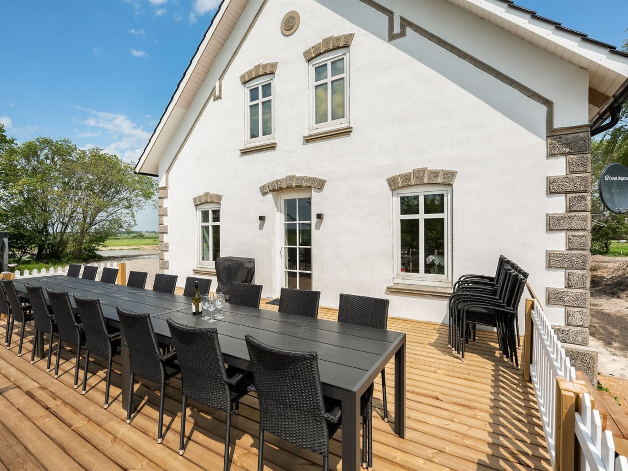 A large outdoor deck with a long dining table and chairs, a covered barbecue area, and a white building with stone accents and multiple windows.