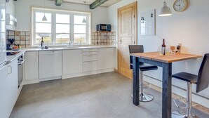 Cocina moderna con alacenas blancas, una mesa de comedor de madera y un taburete de bar. Se ve una ventana con vistas al exterior, un reloj en la pared y un microondas en la encimera.