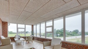 Un salón con mobiliario de mimbre, una mesa de centro de vidrio con fruta, y ventanas grandes que ofrecen vistas al campo.