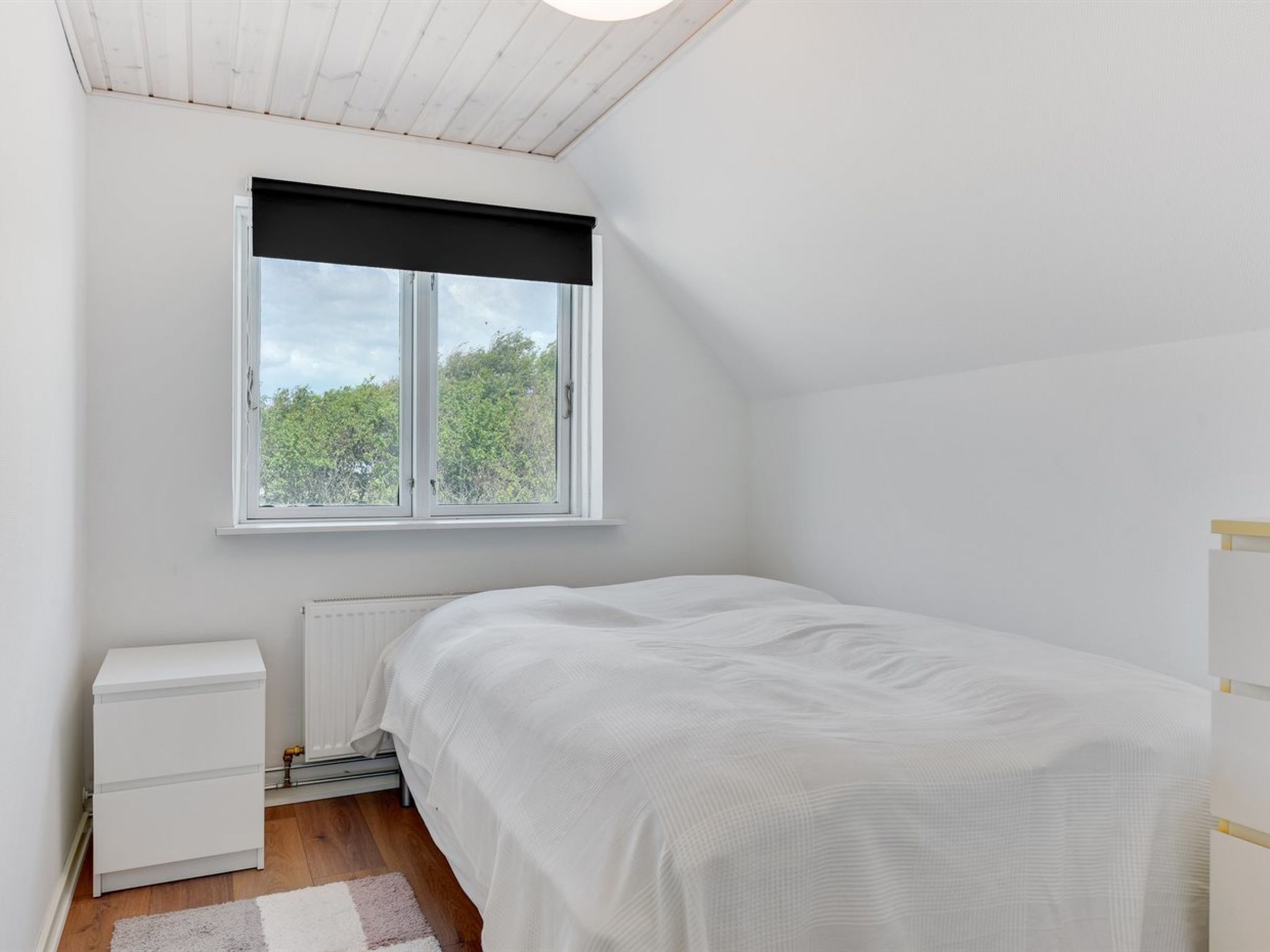 Une petite chambre à coucher blanche, avec un lit simple, un radiateur, une table de chevet blanche et une fenêtre avec un store noir.