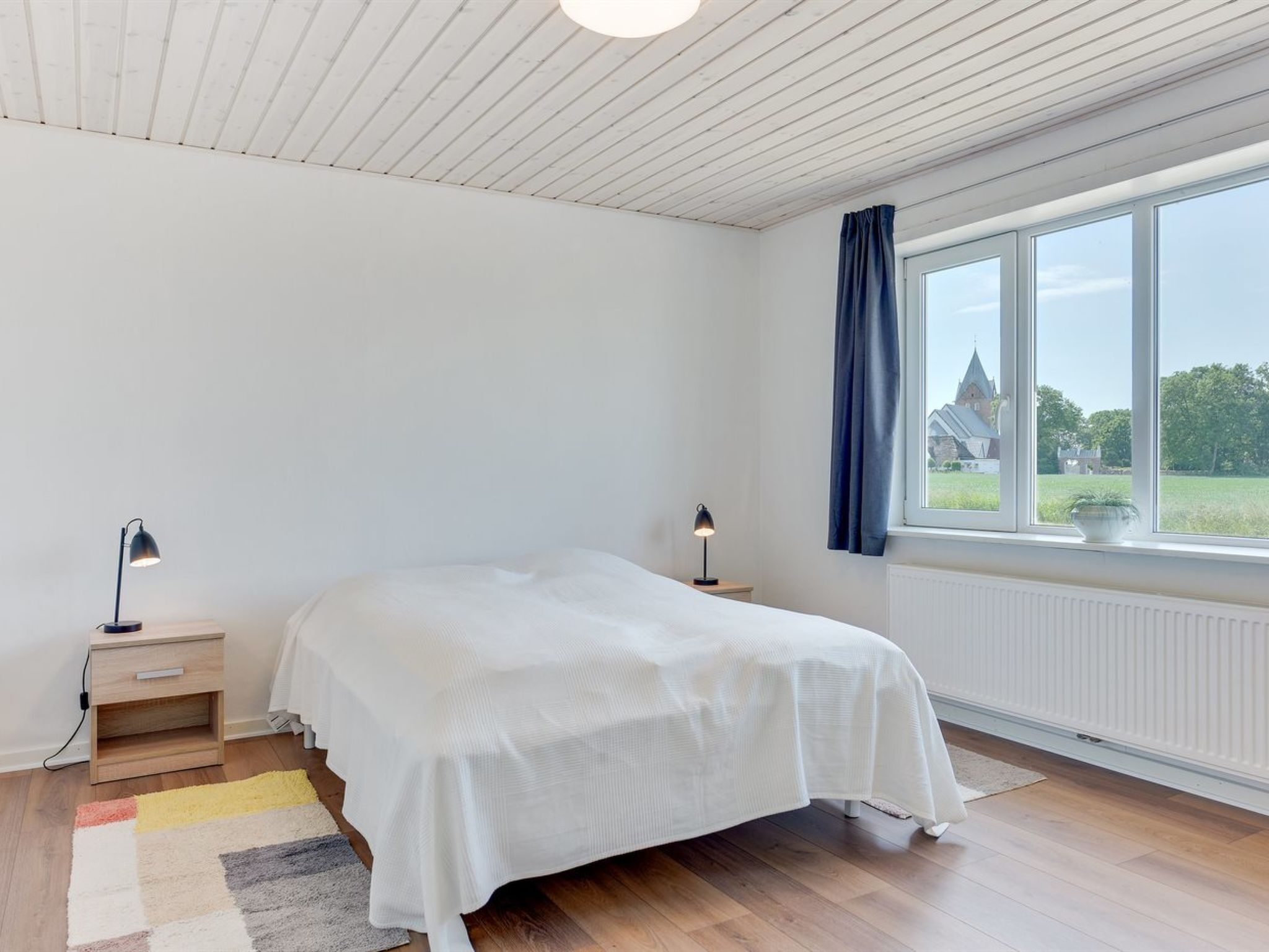 A bedroom with a bed, a nightstand, a lamp, a window with curtains, and a view of a building outside.