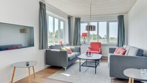 Ein modernes Wohnzimmer mit Fernseher, grauen Sofas, einem Couchtisch und großen Fenstern mit Vorhängen.