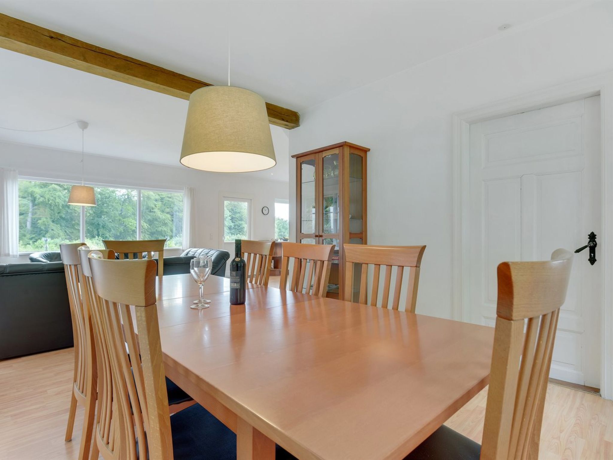 Comedor con mesa y sillas de madera, una lámpara colgante y vistas al exterior a través de amplios ventanales.