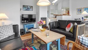 A modern living room with a flat-screen TV, a blue cabinet, a black leather sofa, a wooden coffee table, and a dining area with a white table and chairs.