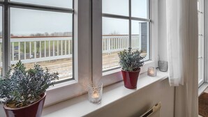 A window with a view of a white fence and a train track. There are two potted plants on the windowsill and two lit candle holders.