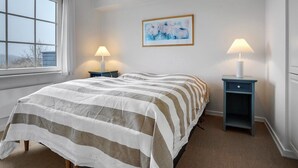 A neatly arranged bedroom with a large bed, two bedside tables with lamps, a framed abstract painting, and a window with curtains.