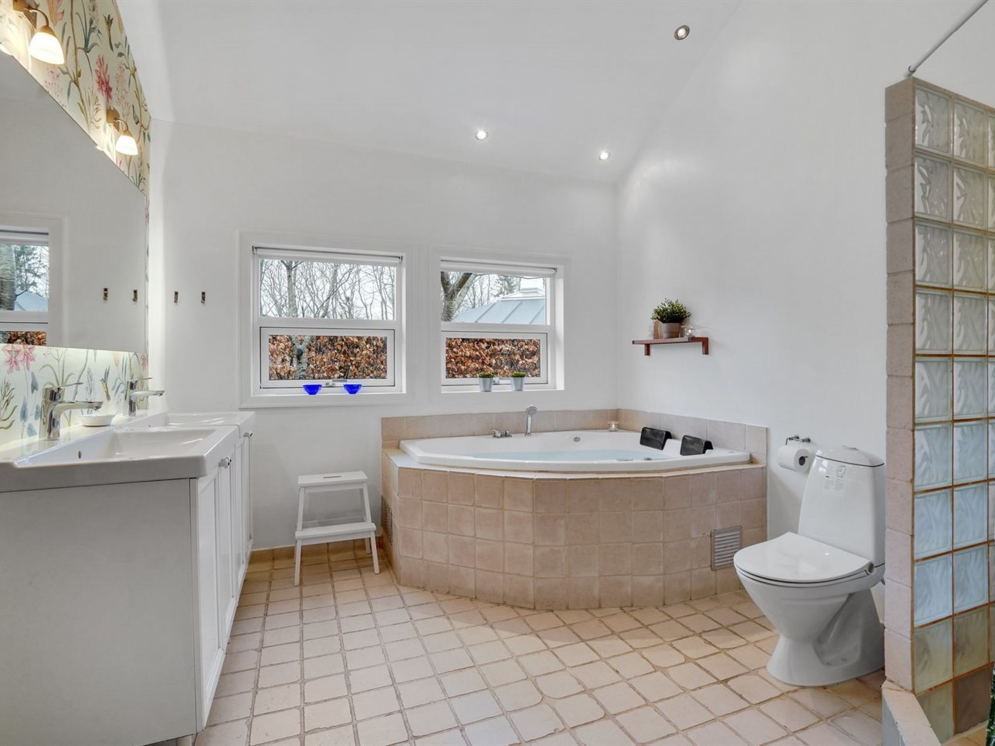 A modern bathroom with a large bathtub, a toilet, and a sink. There are two windows with decorative tiles, a small stool, and a shelf with a plant.