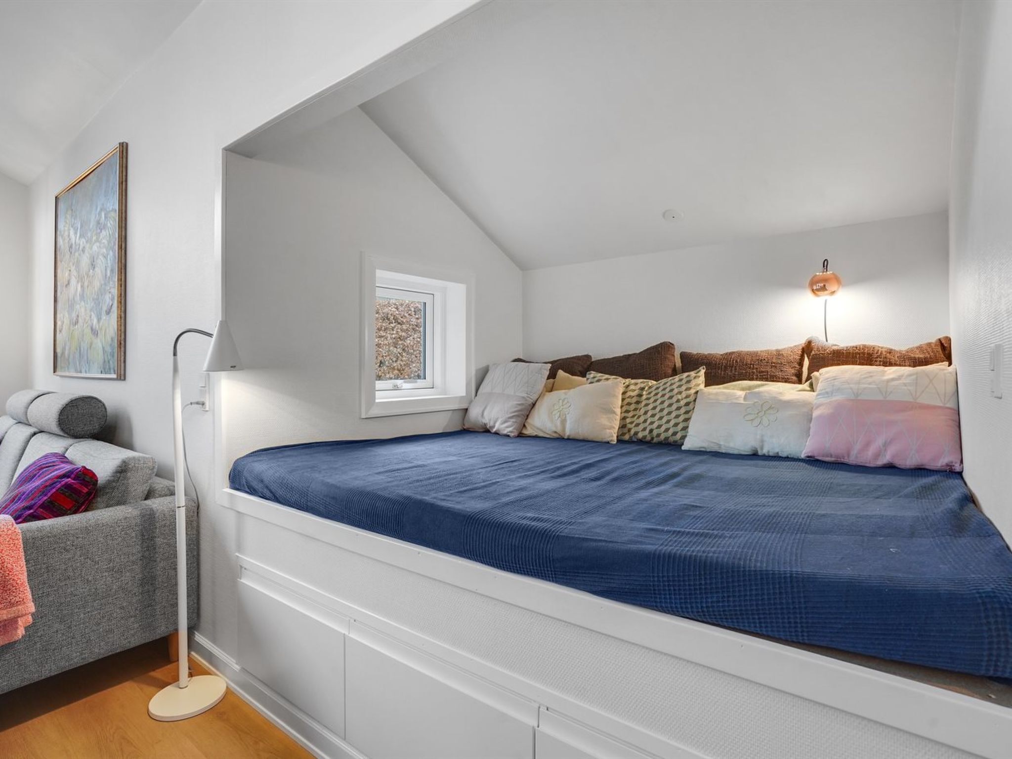 A bedroom with a sloped ceiling, a bed with multiple pillows, a bedside lamp, a framed picture on the wall, and a floor lamp beside a couch.